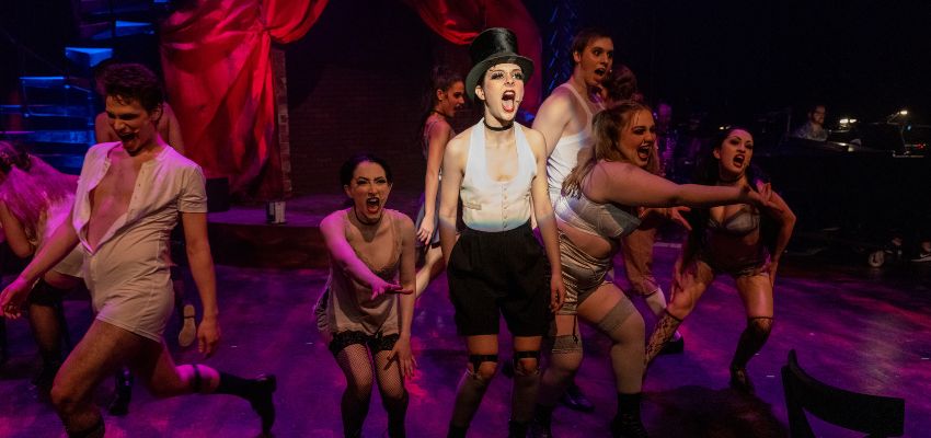 A cabaret MC stands center surrounded by scantly clad dancers.