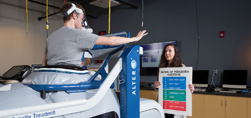 Kinesiology graduate student works with another graduate student on the anti-gravity treadmill in the Exercise Physiology Lab in the Professional Studies building.