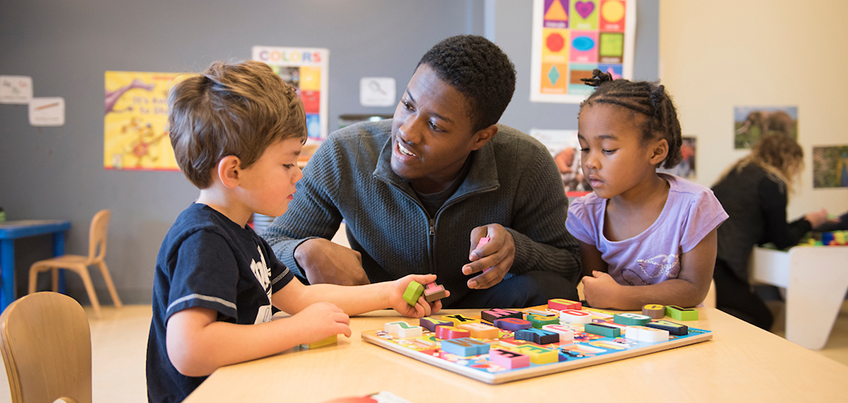 A male student teacher works with two young children in class to put together a puzzle