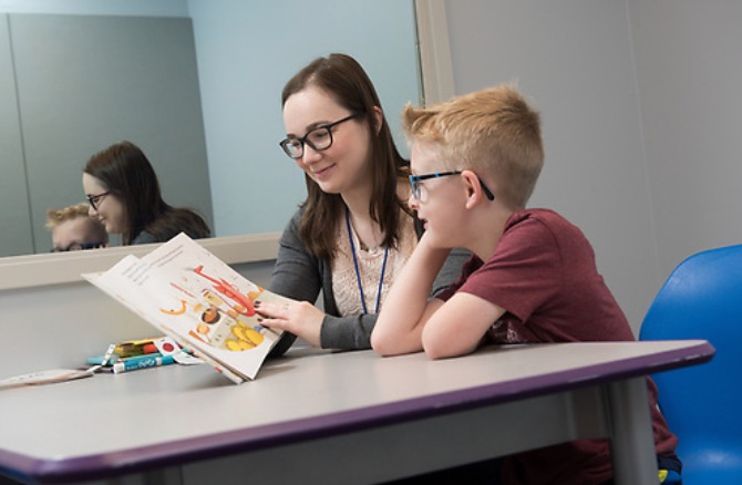 An undergraduate student sitting at a table with a boy reading a book